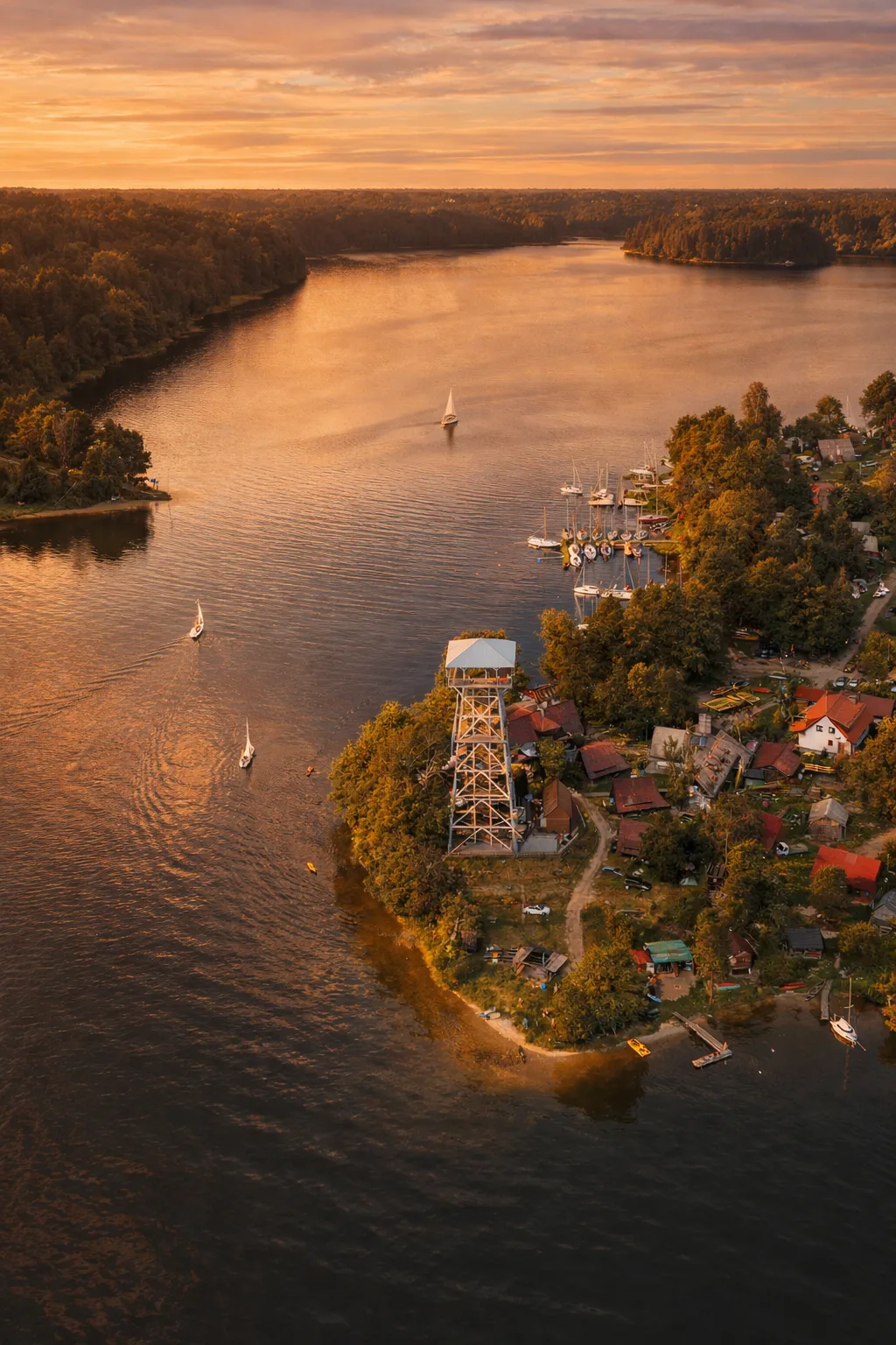Widok z lotu ptaka na wieżę widokową i przystań w Wdzydzach Kiszewskich podczas złotej godziny – półwysep otoczony jeziorem, żaglówki na wodzie i ciepłe, pomarańczowe światło zachodzącego słońca nad Kaszubami. Wdzydze Kiszewskie