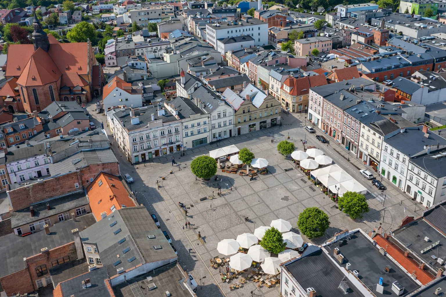 Rynek miasta z lotu ptaka – zabytkowa zabudowa wokół placu, czerwony kościół z wieżą, kamienice w pastelowych kolorach oraz ogródki kawiarniane z białymi parasolami widziane w ostrym, słonecznym świetle.
