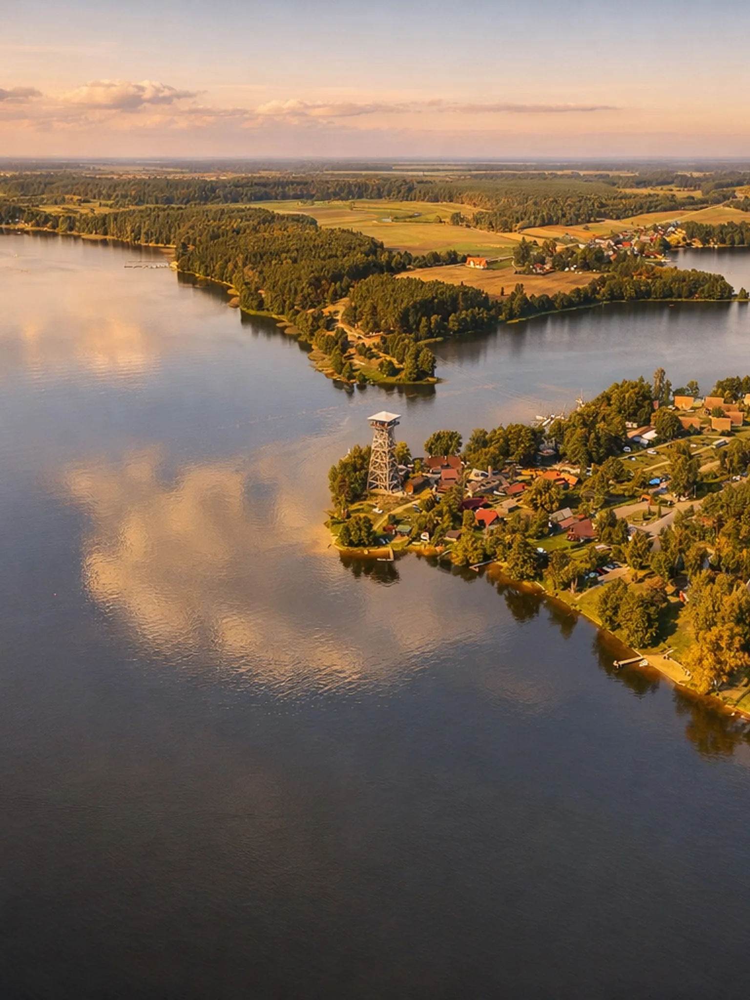 Lotnicze zdjęcie jeziora o złotej godzinie – spokojna tafla wody półwysep z wieżą widokową, domkami i lasem, w tle pola i niewielka miejscowość w ciepłym, delikatnym świetle zachodzącego słońca.