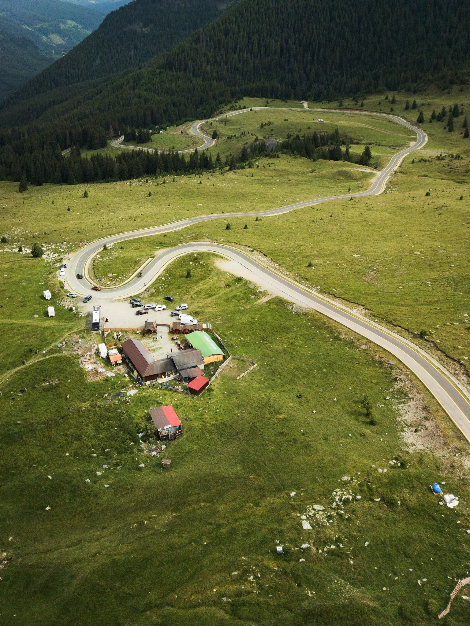 Serpentyny Trasy Transalpina i zabudowania przydrożne widziane z drona na tle lasów i górskich łąk.