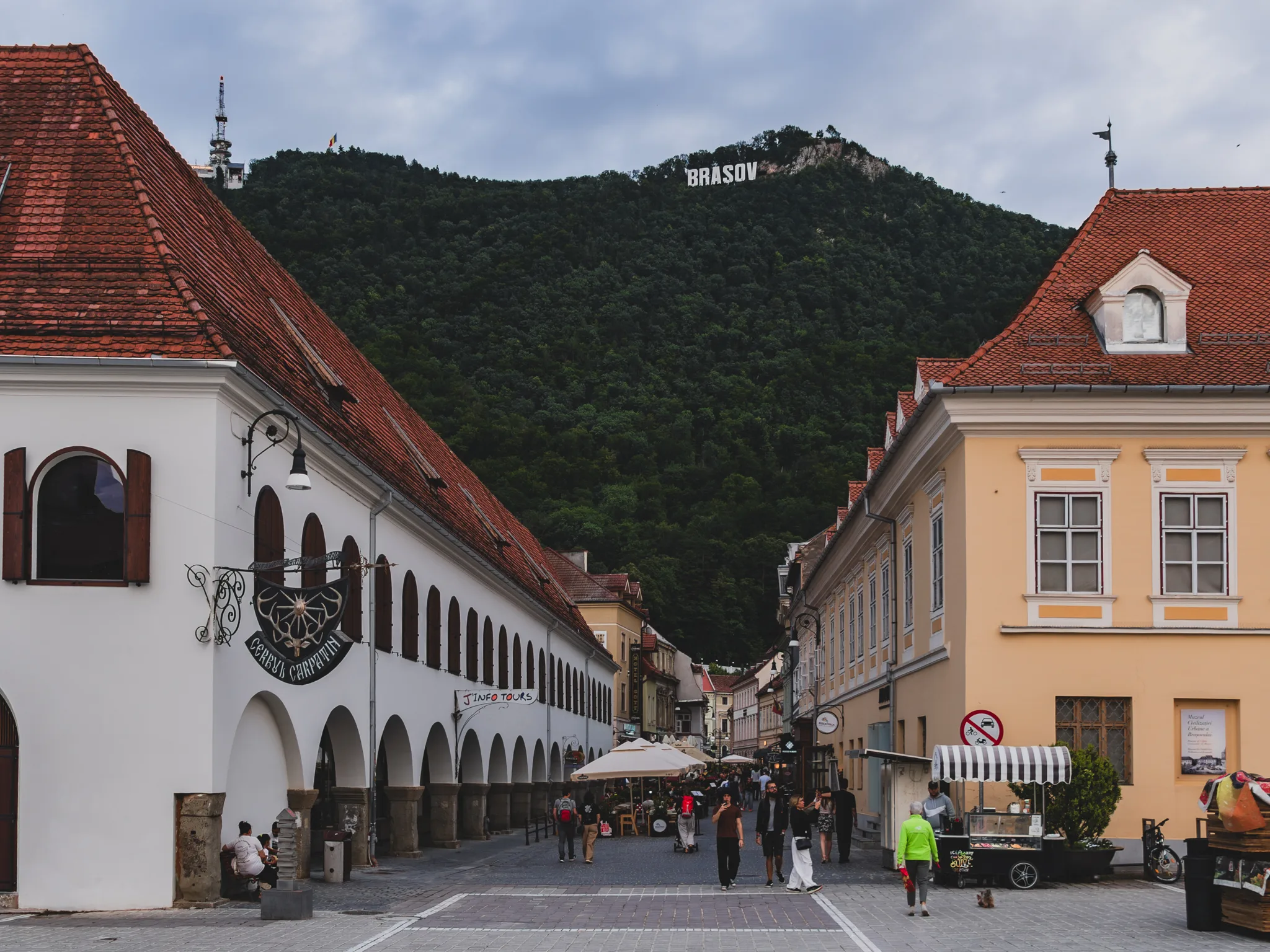 Ulica na Starym Mieście w Braszowie z zabytkowymi kamienicami, arkadami i wzgórzem Tâmpa z napisem BRASOV w tle.