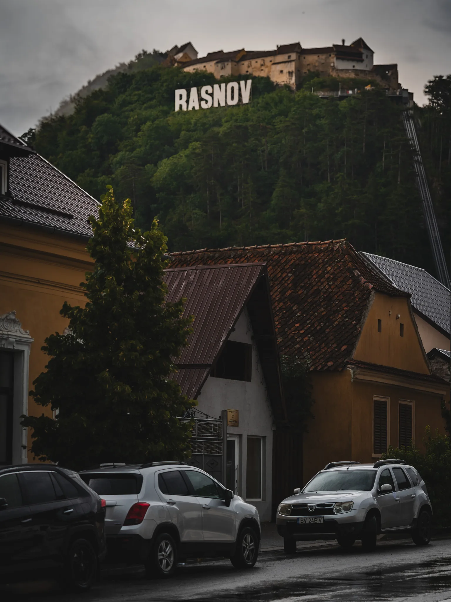 Twierdza Râșnov na wzgórzu nad zabudową miasta, z napisem RASNOV i samochodami na pierwszym planie.