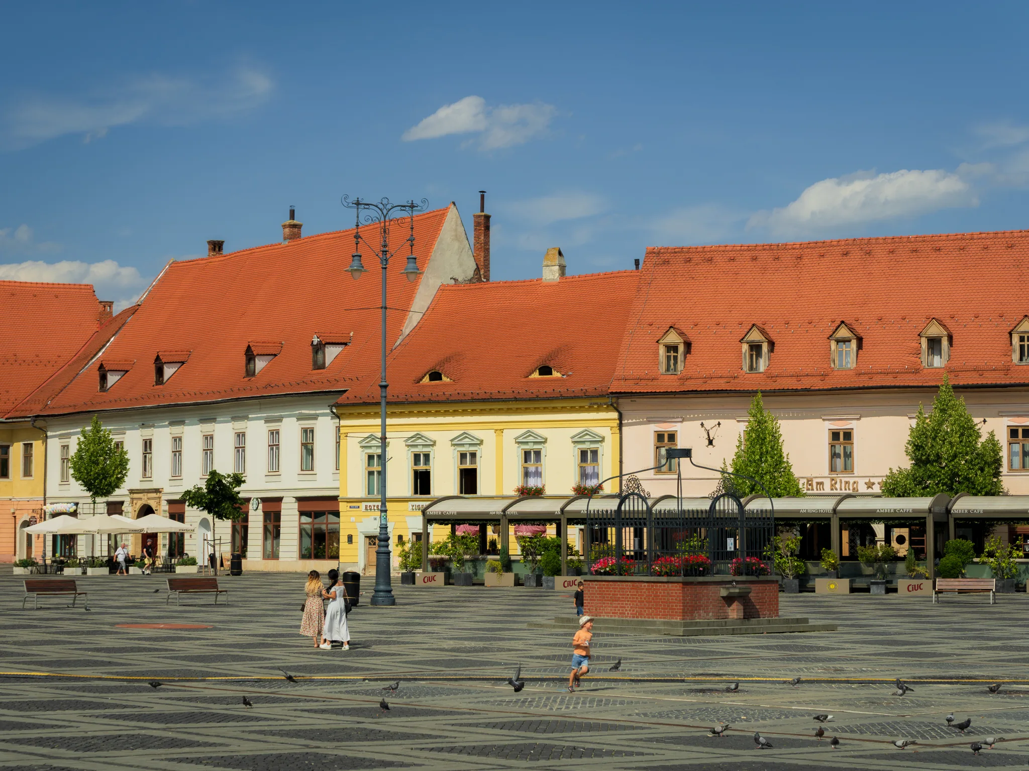 Kolorowe kamienice z czerwonymi dachami przy dużym rynku w Sybinie, na placu spacerują ludzie i gołębie, a nad miastem widać błękitne niebo z chmurami.