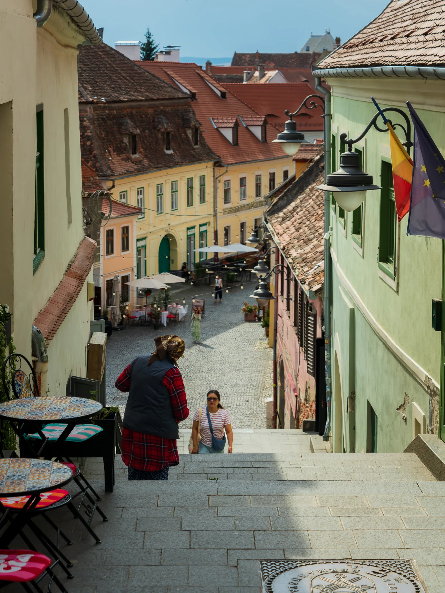 Pasaż Schodów w Sybinie z widokiem na kolorowe kamienice Dolnego Miasta, brukowaną uliczkę oraz flagi Rumunii i Unii Europej.