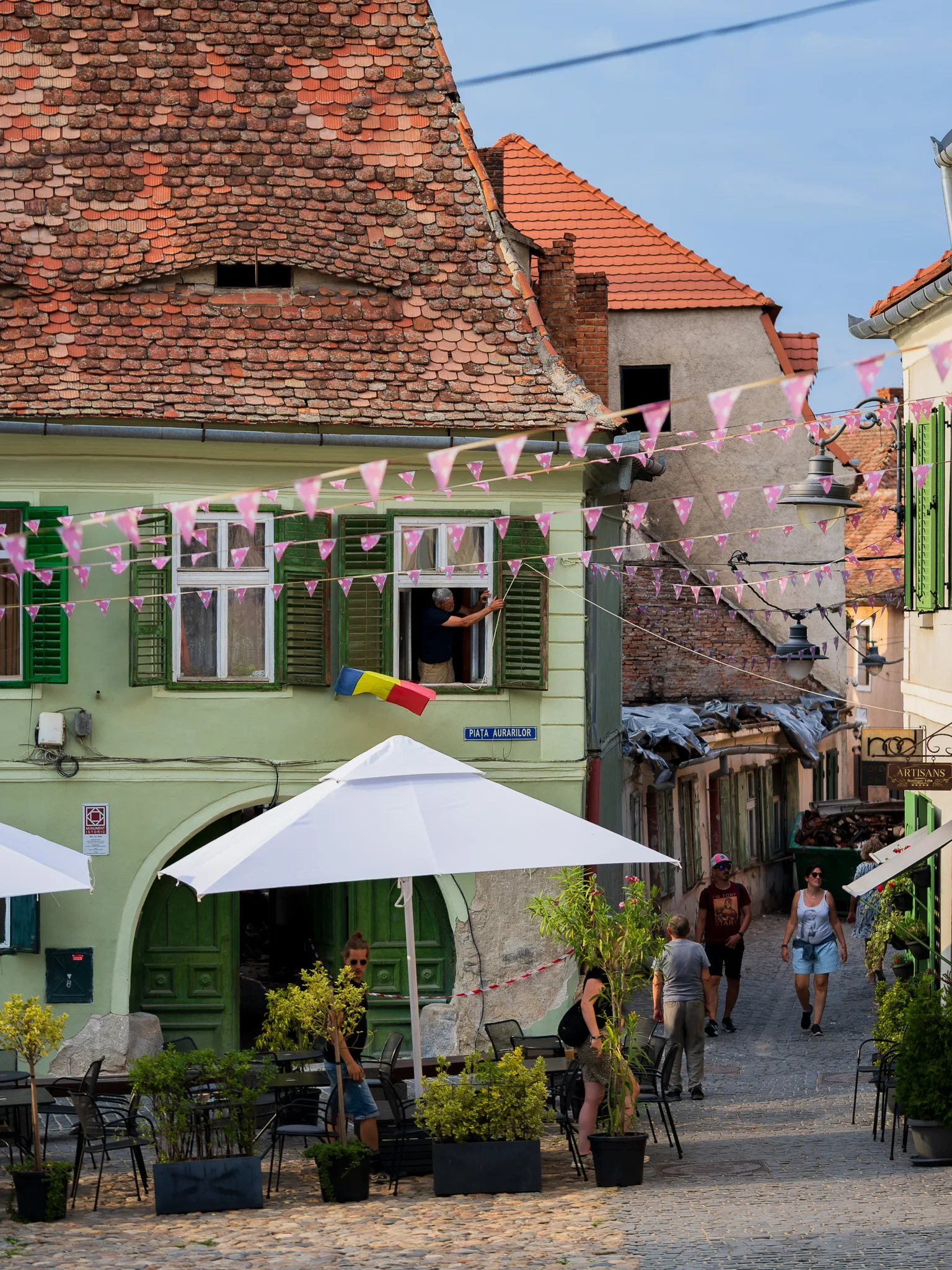 Kolorowe kamienice i brukowana uliczka na starówce w Sibinie z rozwieszonymi flagami, ogródkami restauracyjnymi i przechodniami.