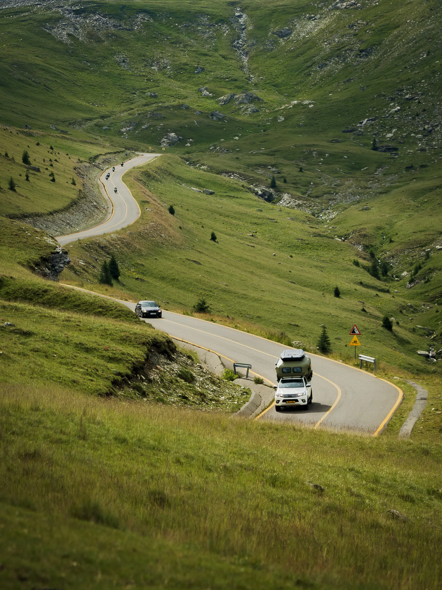 Samochody i motocykle jadące krętą Trasą Transalpina przez zielone zbocza gór Parâng w Rumunii.