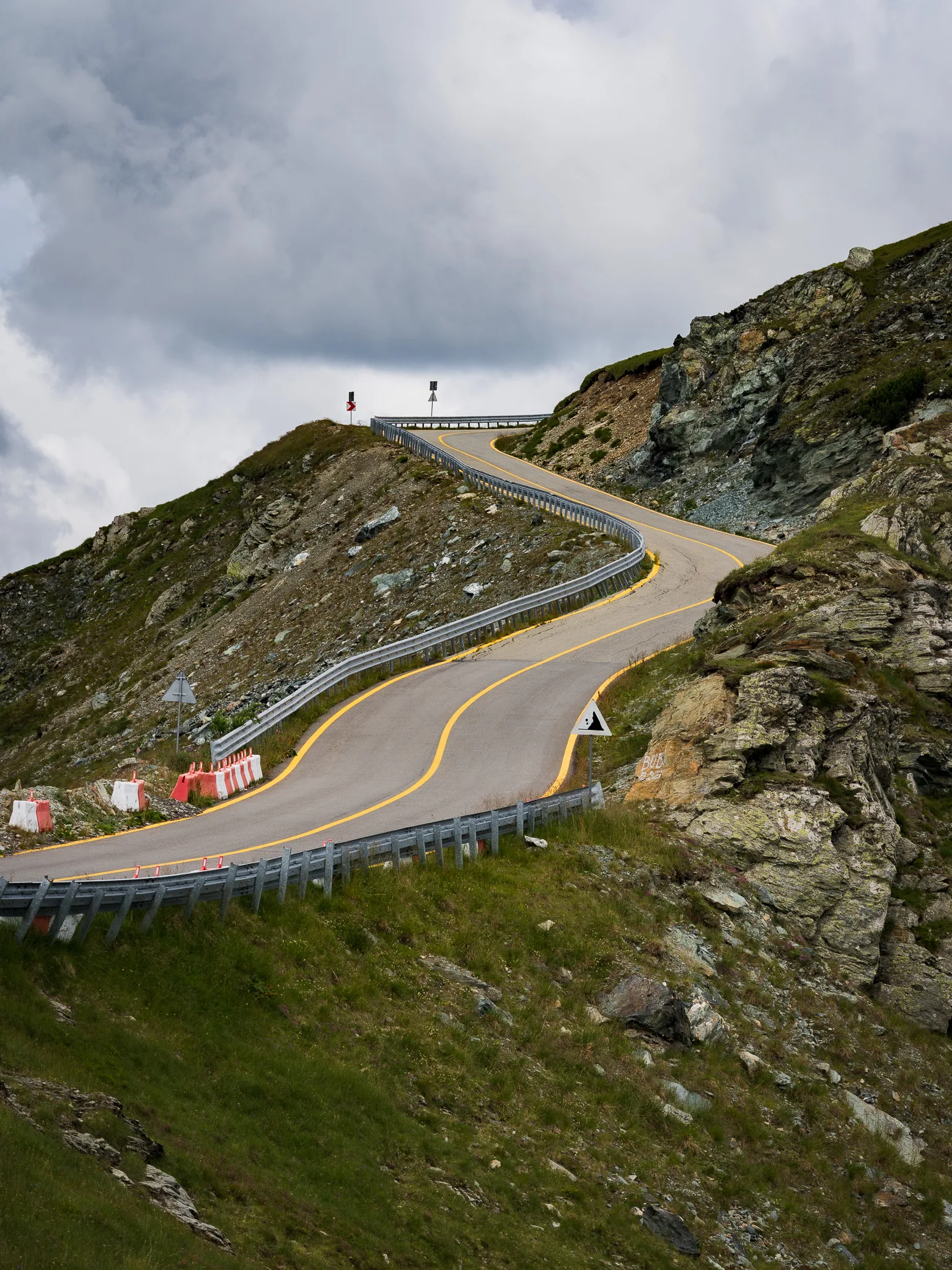 Transalpina prowadząca przez skaliste, wysokogórskie zbocze, z krętą drogą znikającą za granią pod ciężkimi chmurami.