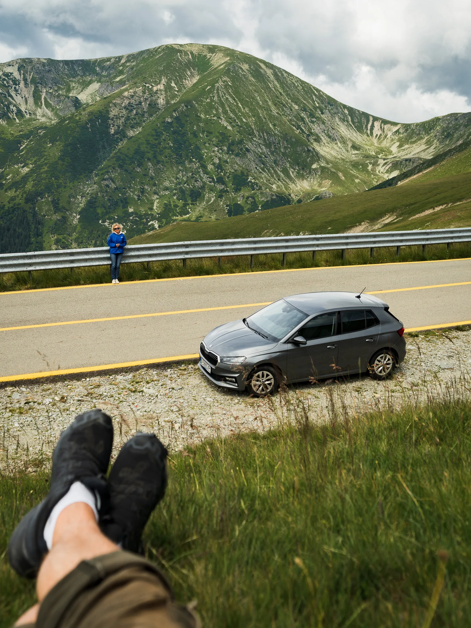 Samochód zaparkowany przy Trasie Transalpina, kobieta przy barierce i panorama gór Parâng w tle.