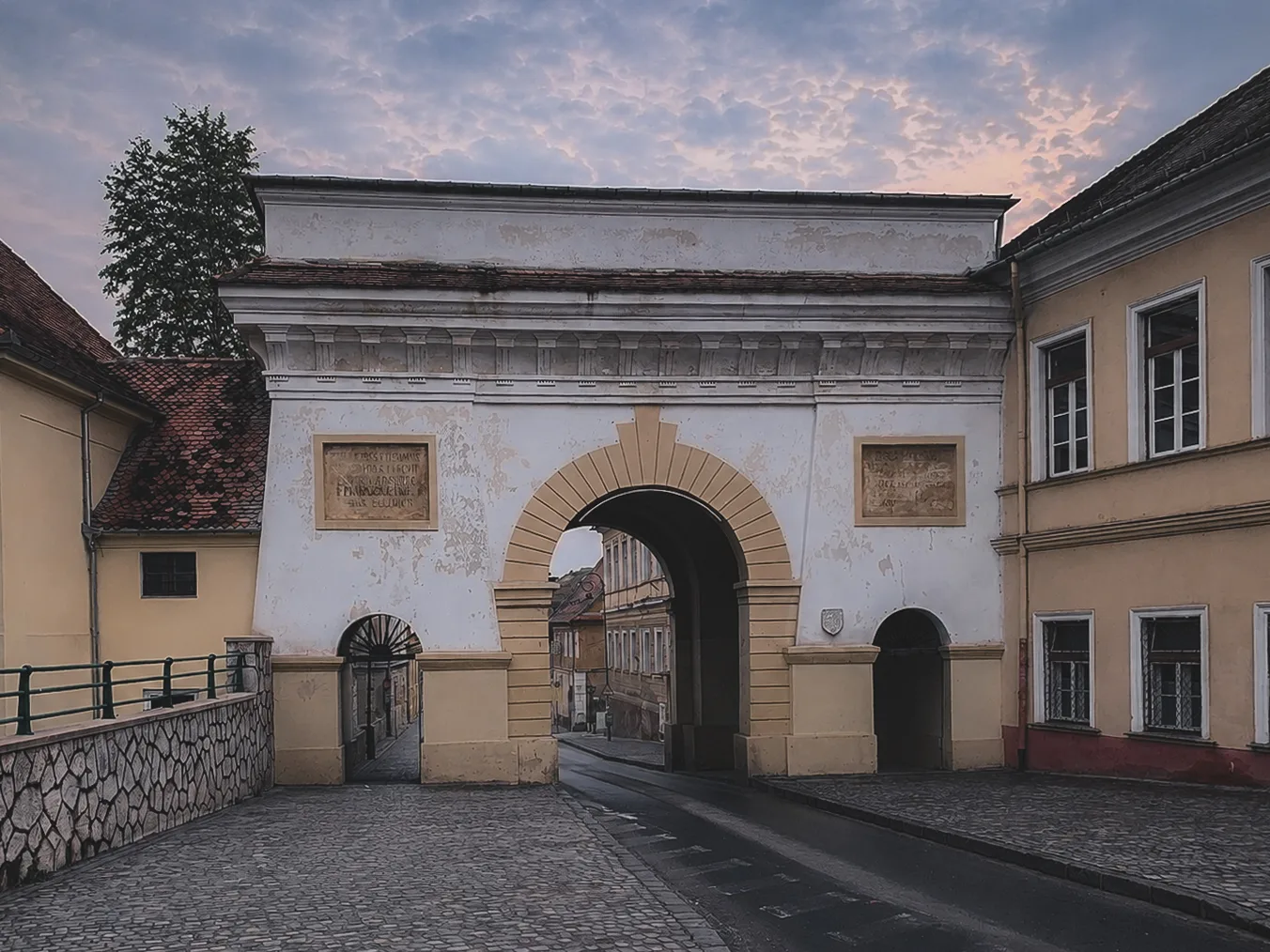 Brama miejska w Braszowie o zmierzchu, z pustą brukowaną ulicą, łagodnym światłem i chmurami w odcieniach różu i błękitu nad zabytkową zabudową.