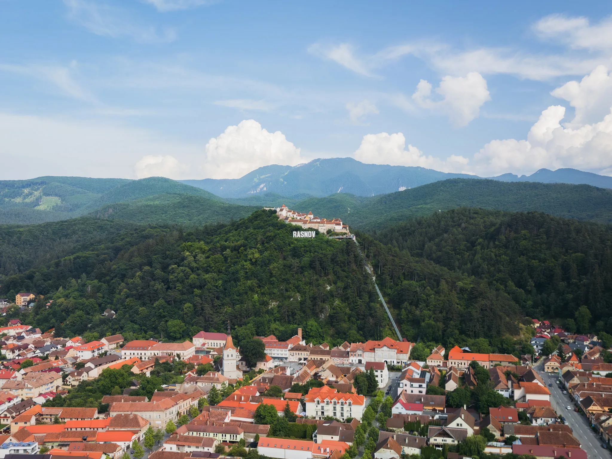 Widok z drona na Râșnov i twierdzę Râșnov położoną na zalesionym wzgórzu, z górami w tle.