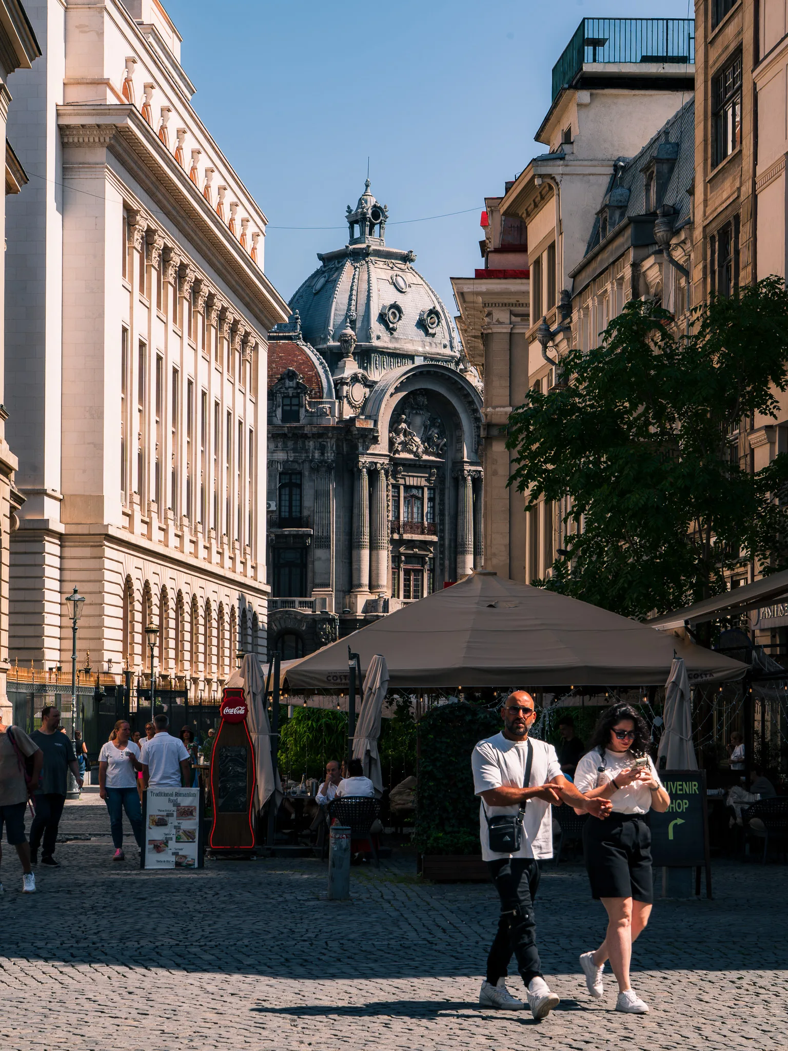 Pałac CEC na końcu uliczki Starego Miasta w Bukareszcie, z ogródkami restauracyjnymi i przechodniami.