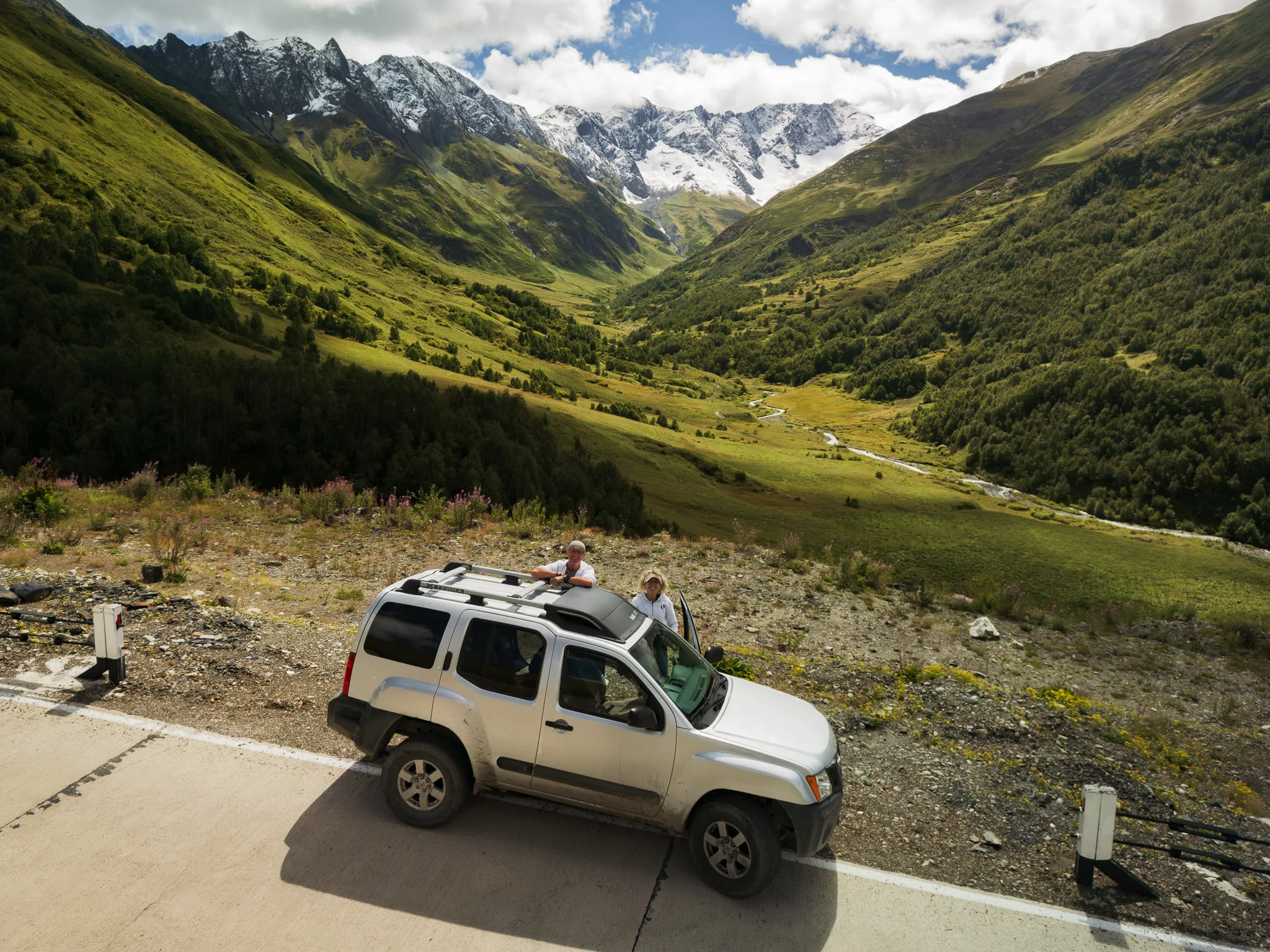 Nissan Xterra 4x4 z parą podróżników na górskiej drodze w Gruzji, z zieloną doliną i ośnieżonymi szczytami Kaukazu w tle.
