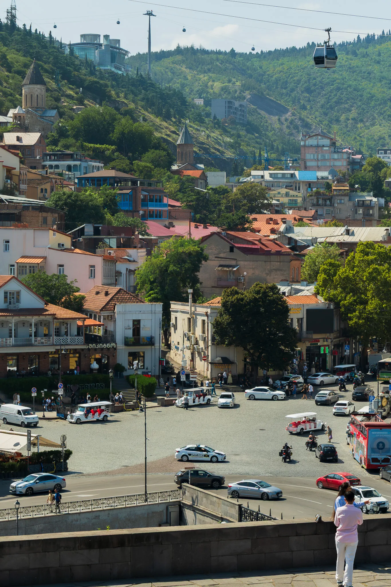 Panorama Starego Miasta w Tbilisi z kolorową zabudową, cerkwiami, ruchem ulicznym i wagonikami kolejki linowej nad miastem.