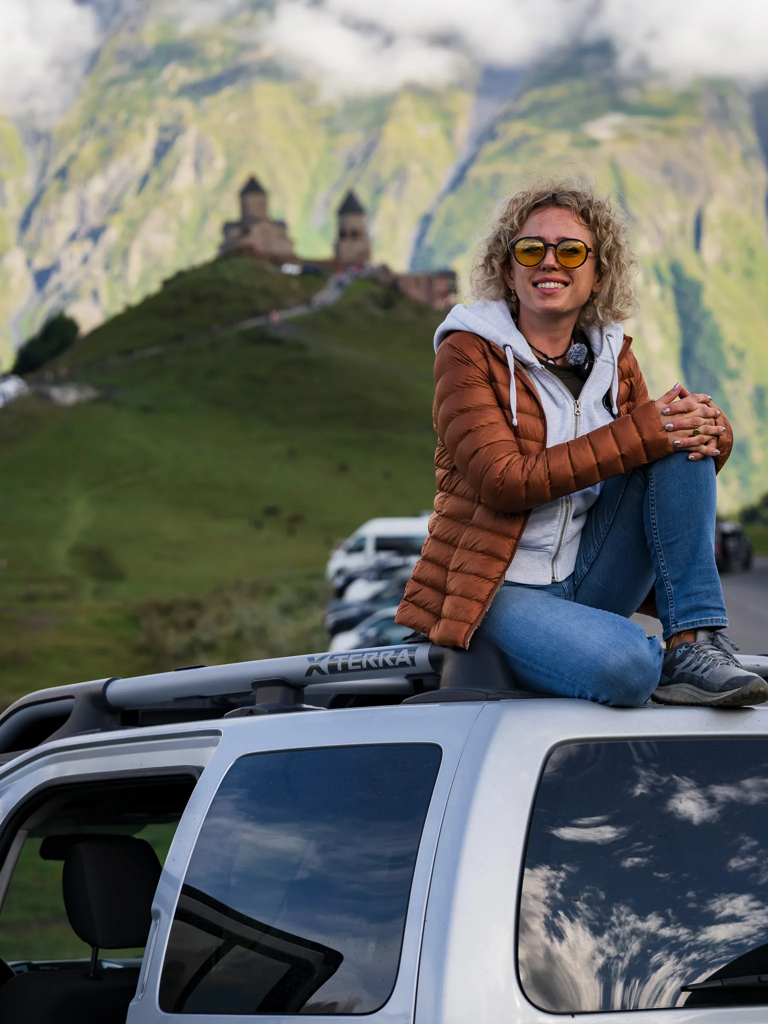 Podróżniczka siedząca na dachu Nissana Xterra 4x4 z cerkwią Cminda Sameba w Gergeti i górami Kaukazu w tle. Gruzja samochodem.