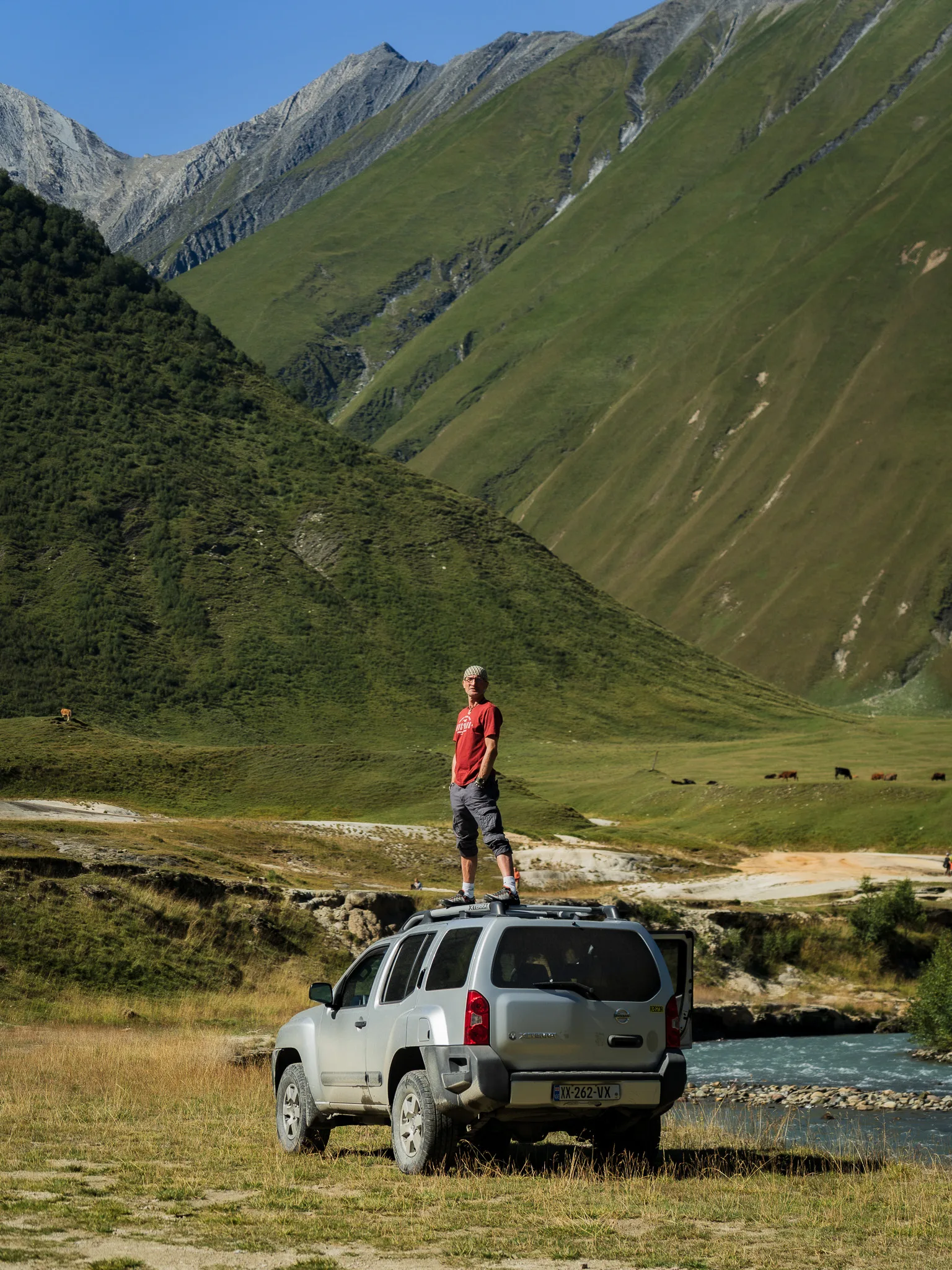 Podróżnik stojący na dachu Nissana Xterra 4x4 w Dolinie Truso w Gruzji, z rzeką i zielonymi zboczami Kaukazu w tle.