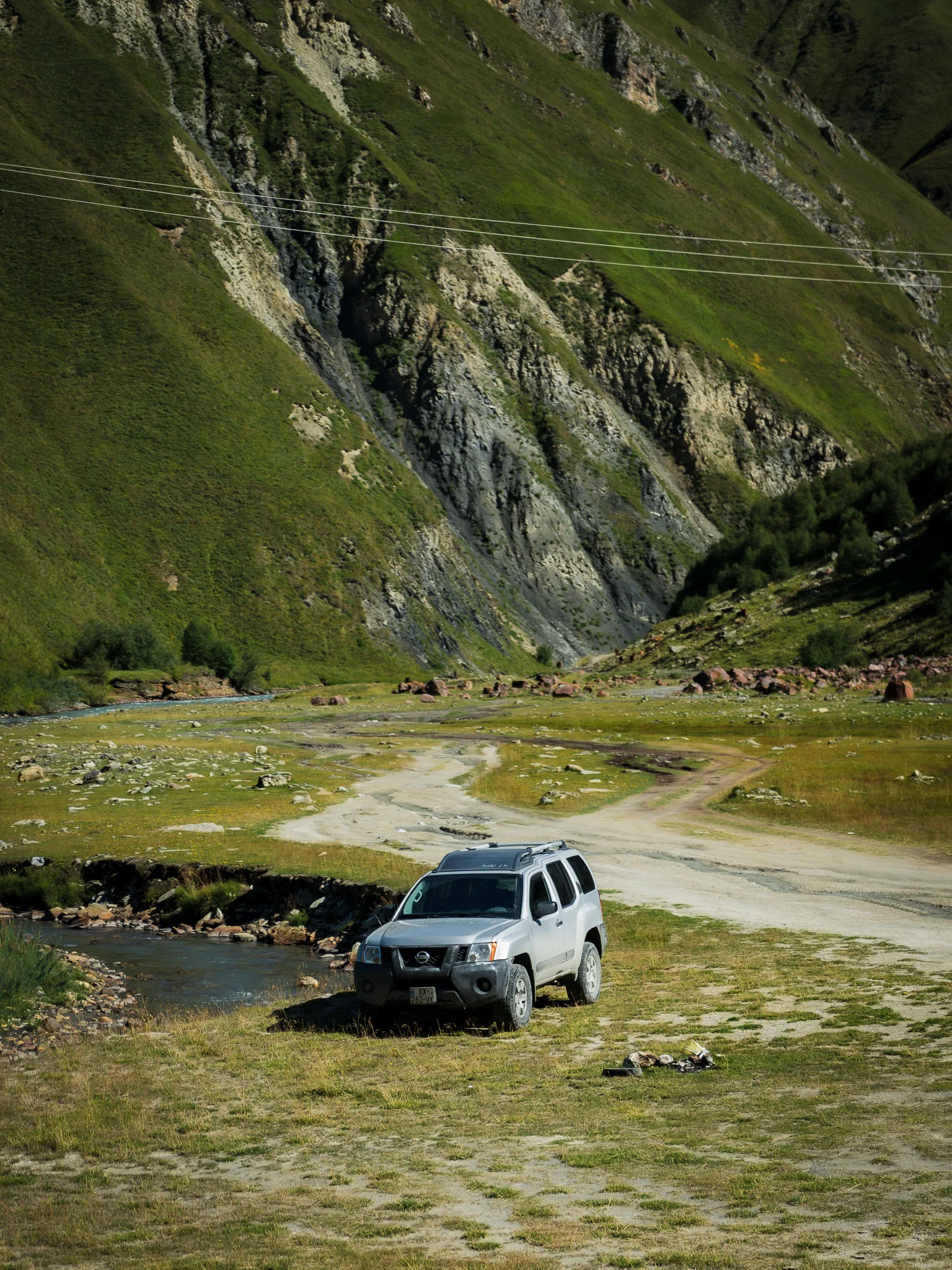 Nissan Xterra 4x4 zaparkowany przy górskiej rzece w Dolinie Truso w Gruzji, na tle zielonych zboczy Kaukazu i szutrowej drogi.