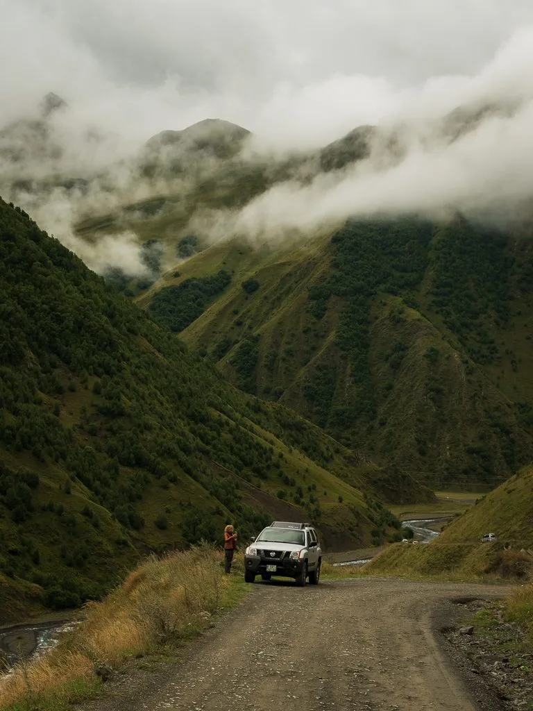 Nissan Xterra 4x4 na szutrowej górskiej drodze w regionie Kazbegi w Gruzji, wśród zielonych zboczy spowitych niskimi chmurami.