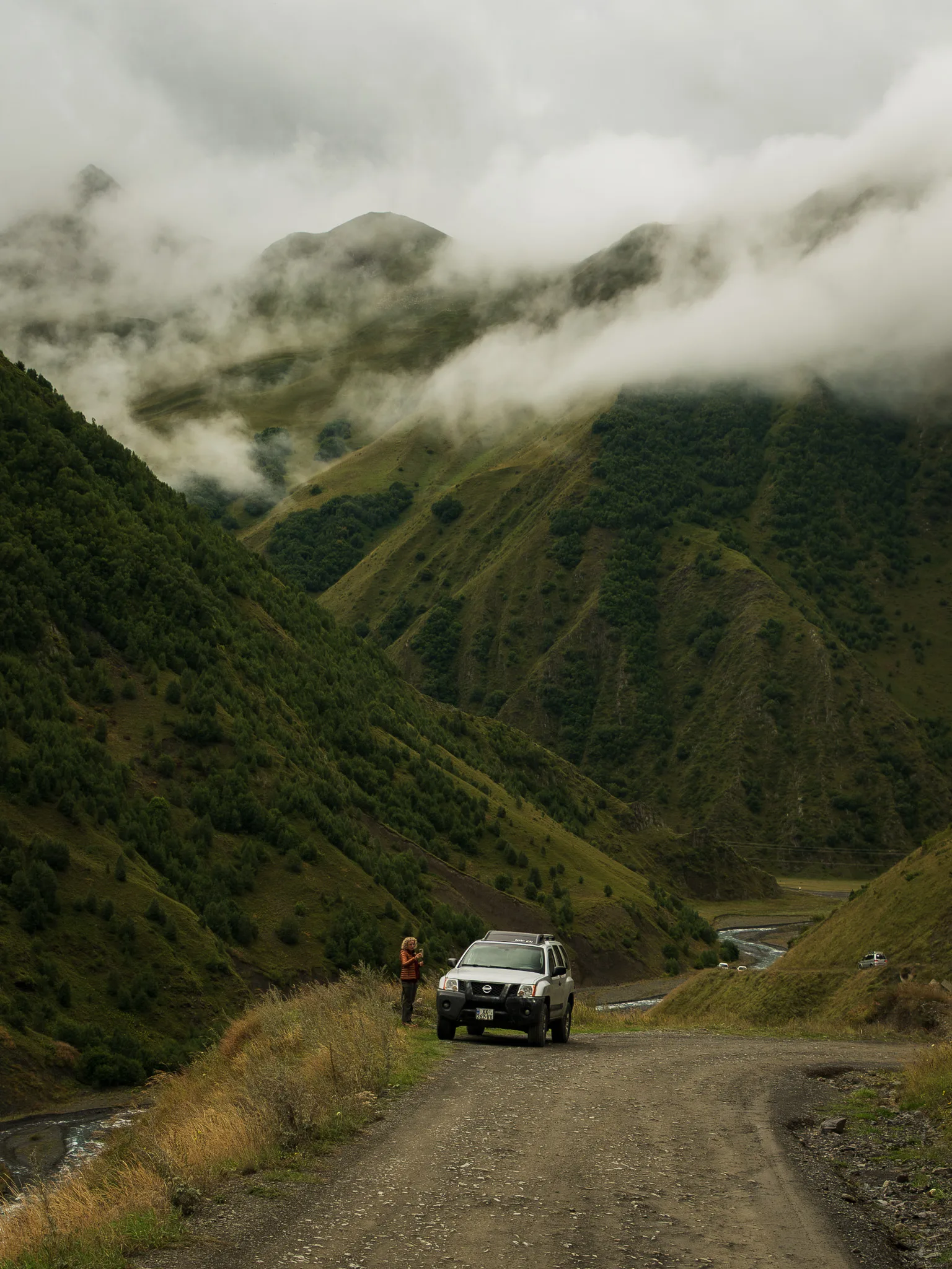 Nissan Xterra 4x4 na szutrowej górskiej drodze w regionie Kazbegi w Gruzji, wśród zielonych zboczy spowitych niskimi chmurami.
