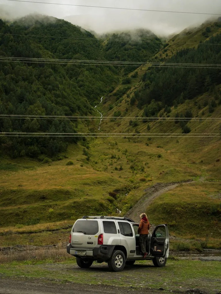 Nissan Xterra 4x4 zaparkowany w górskiej dolinie w Gruzji, z podróżniczką przy aucie i mgłą opadającą na zielone zbocza Kaukazu.