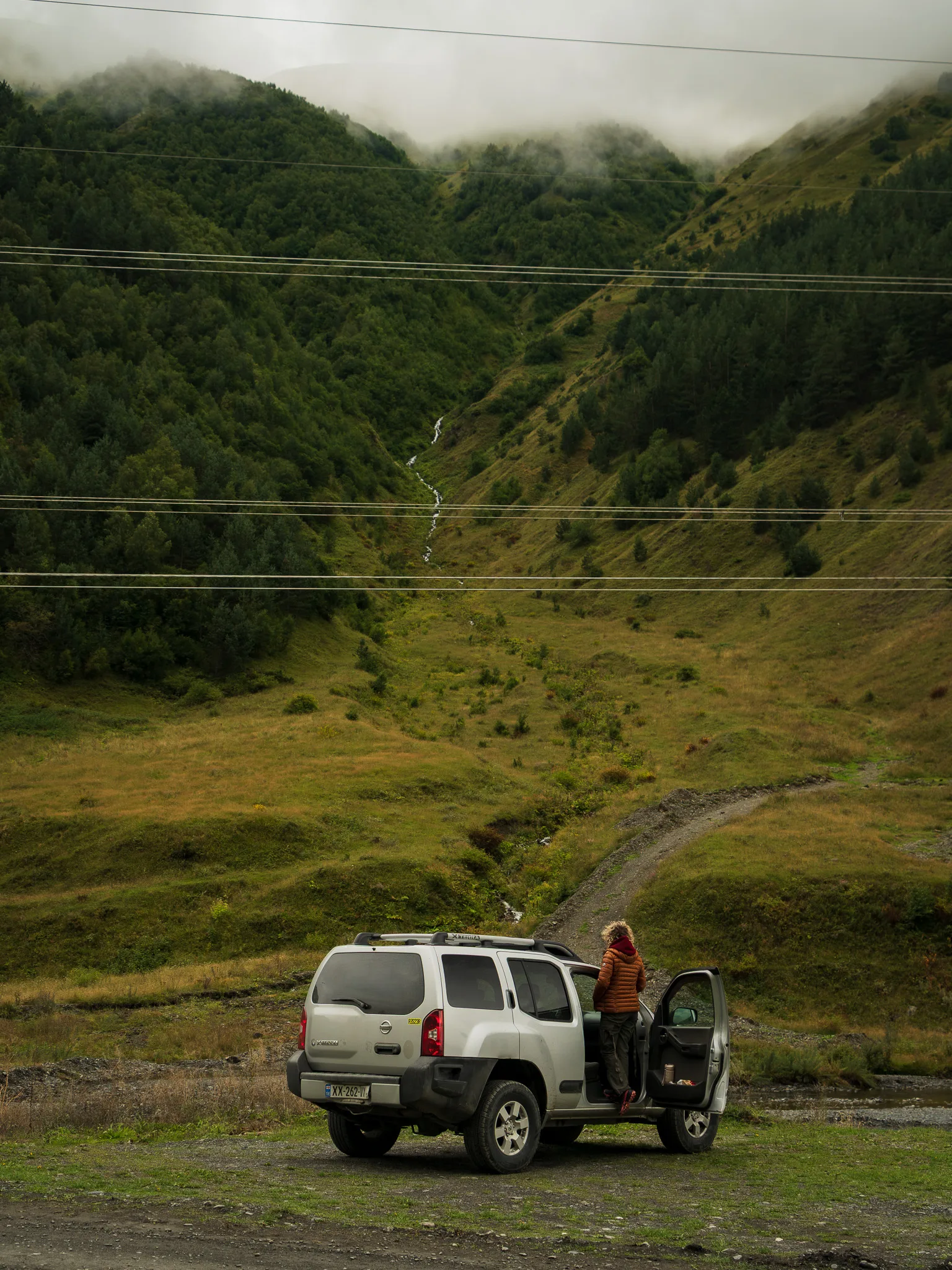 Nissan Xterra 4x4 zaparkowany w górskiej dolinie w Gruzji, z podróżniczką przy aucie i mgłą opadającą na zielone zbocza Kaukazu.